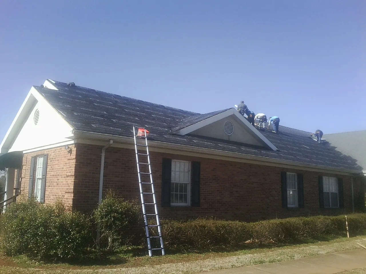 Crew tearing shingles off a brick ranch roof with a ladder set — Emergency Roof Repair work by our crew in Lilesville
