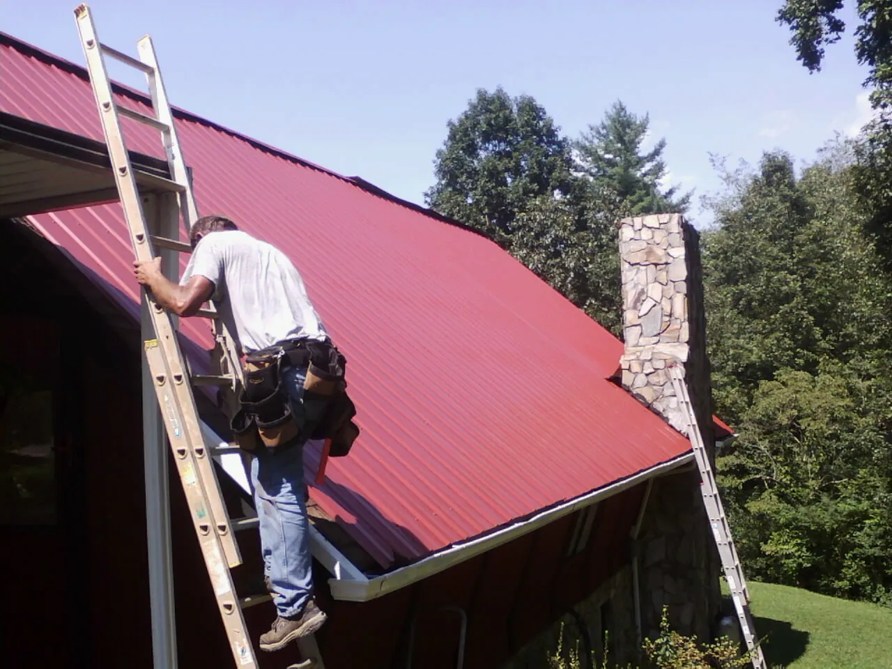 Roofer on a ladder beside a red metal roof and stone chimney — Soffit & Fascia Repair by Roofing Brothers in Lilesville